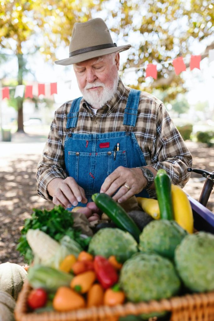Mature man wearing overalls and a hat with a basket full of freshly harvested vegetables.