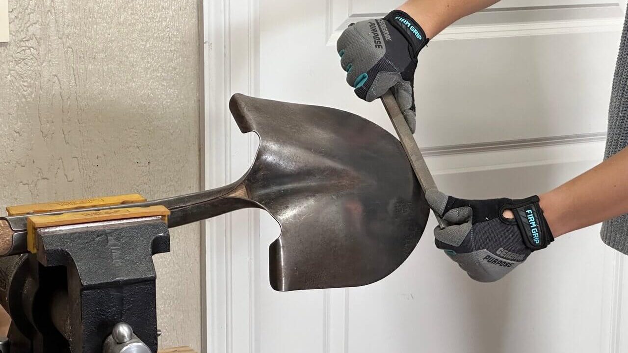 Image of a persons forearms with gloved hands holding a flat file while sharpening the edge of a shovel that is being held in a vice. This is an example how to sharpen and properly winterize your garden tools.