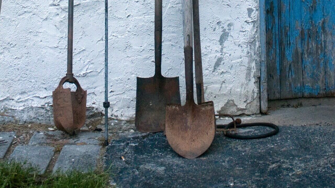 Image of several dirty and rusty garden tools, including 2 shovels and a post hole digger, leaning against the outside of a building. They are examples of garden tools that need to be winterized and stored.